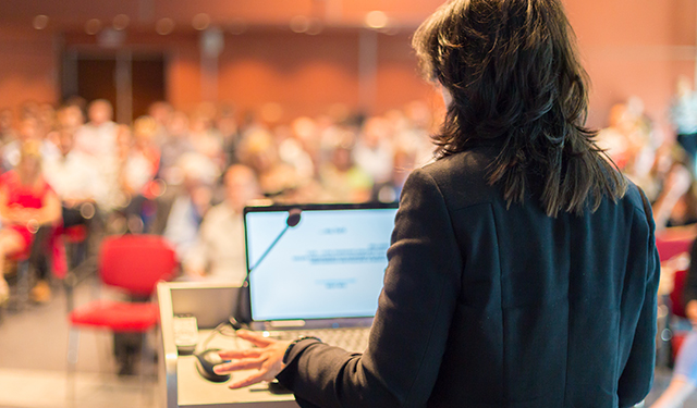 A woman in business attire is giving a lecture to a room full of people. She is silhouetted by the glow of her computer's monitor. 